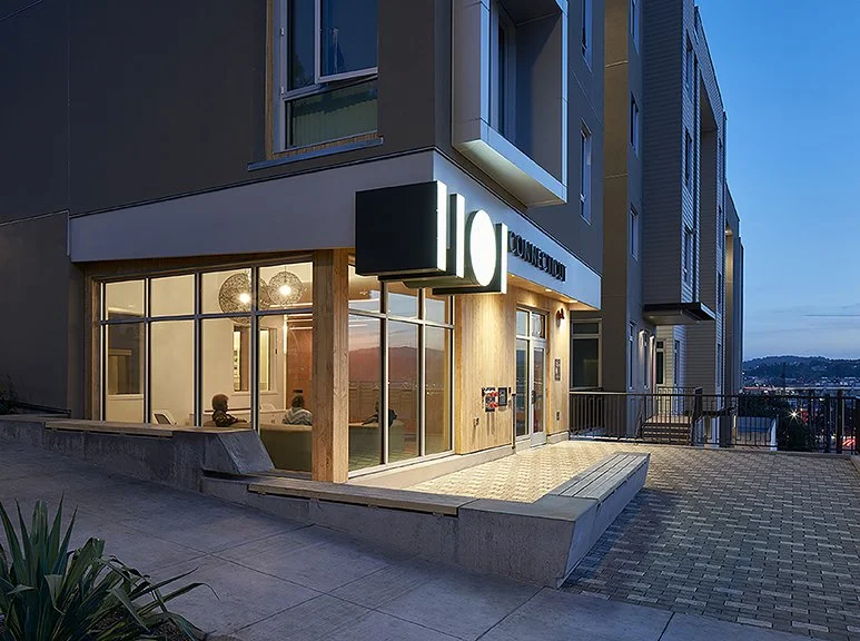 1101 Connecticut entrance at dusk with illuminated signage, wood cladding, and a glass-walled community space.