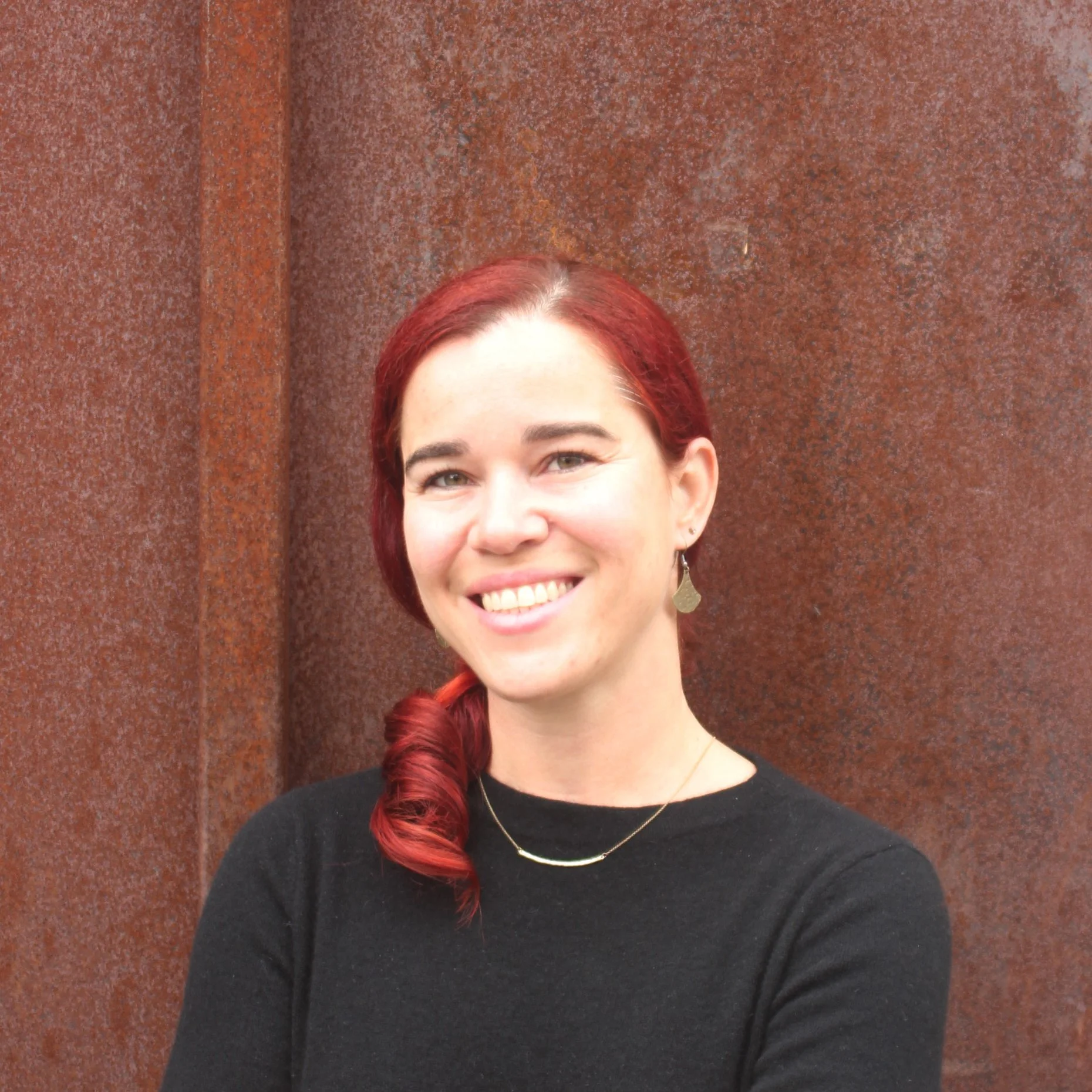 Professional headshot of Leticia Colnago wearing a black top against a rust-colored wall