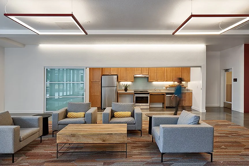 Community lounge with upholstered seating, a wood coffee table, and an open kitchen beyond.