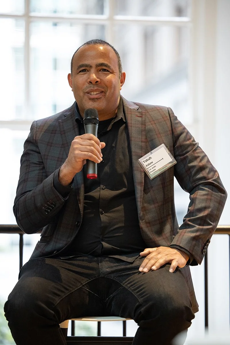 Person seated on a stool speaking into a handheld microphone at an indoor event, wearing a plaid blazer and name badge