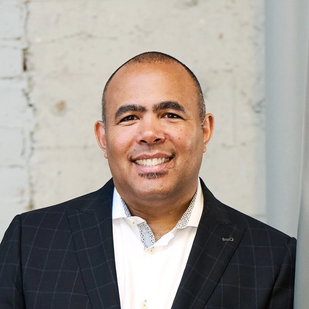 Professional headshot of a man in a dark suit jacket and collared shirt, standing in front of a softly blurred neutral background.
