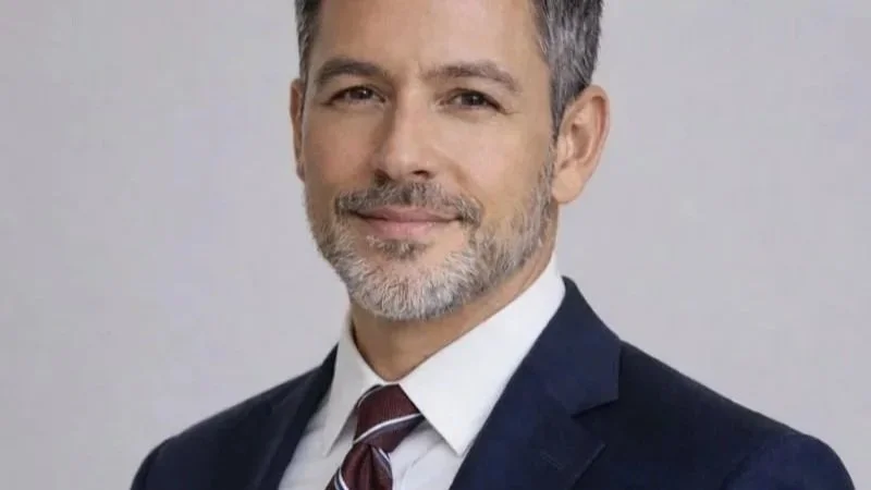 Professional middle-aged man with gray hair and beard, wearing a dark suit, white shirt, and maroon tie, smiling against a plain light gray background.