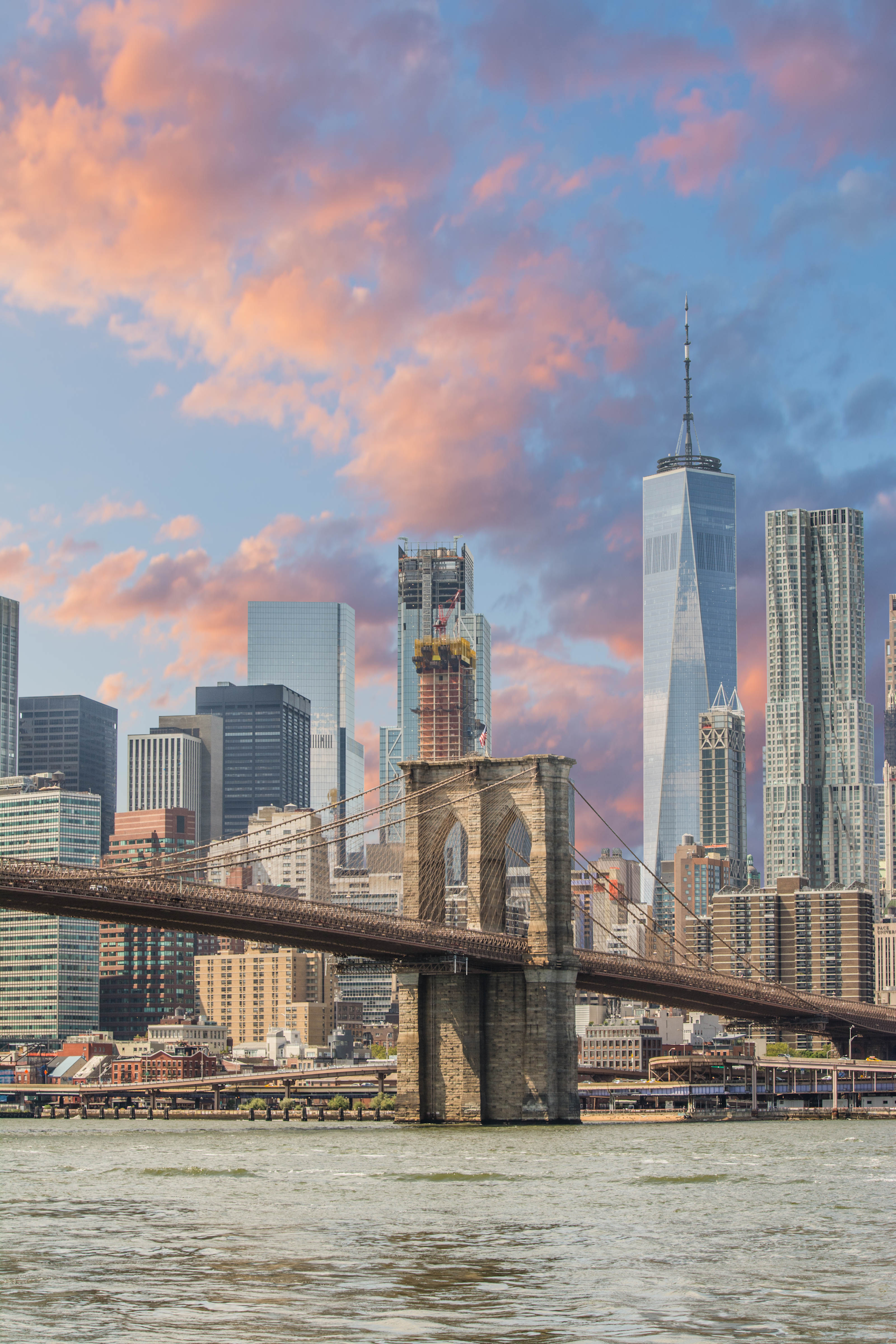 View of New York City skyline showing Brooklyn Bridge and One World Trade Center during sunset with colorful clouds.