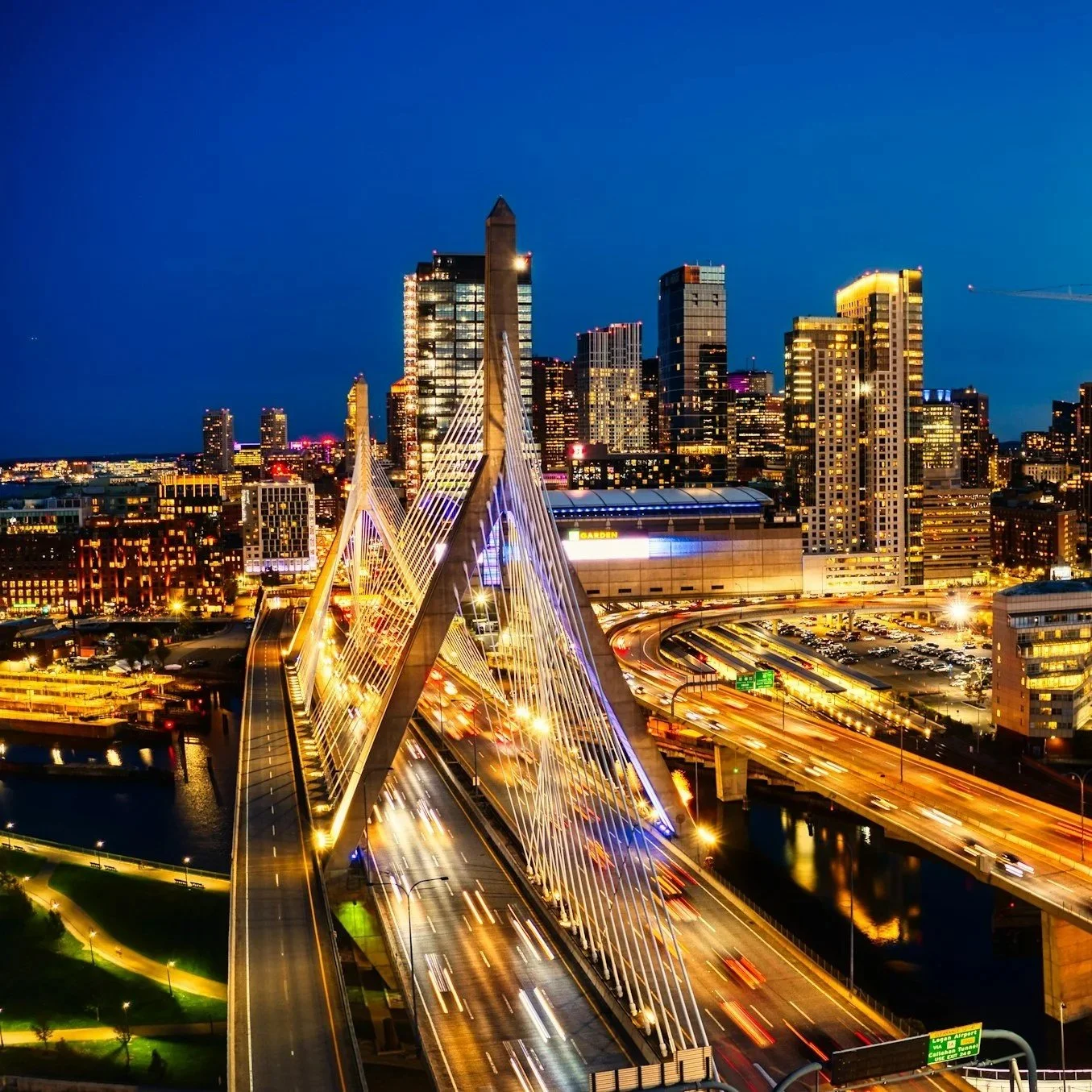 Nighttime cityscape of San Francisco with the Bay Bridge illuminated, showing the skyline with tall buildings and busy roads with light trails from moving vehicles.