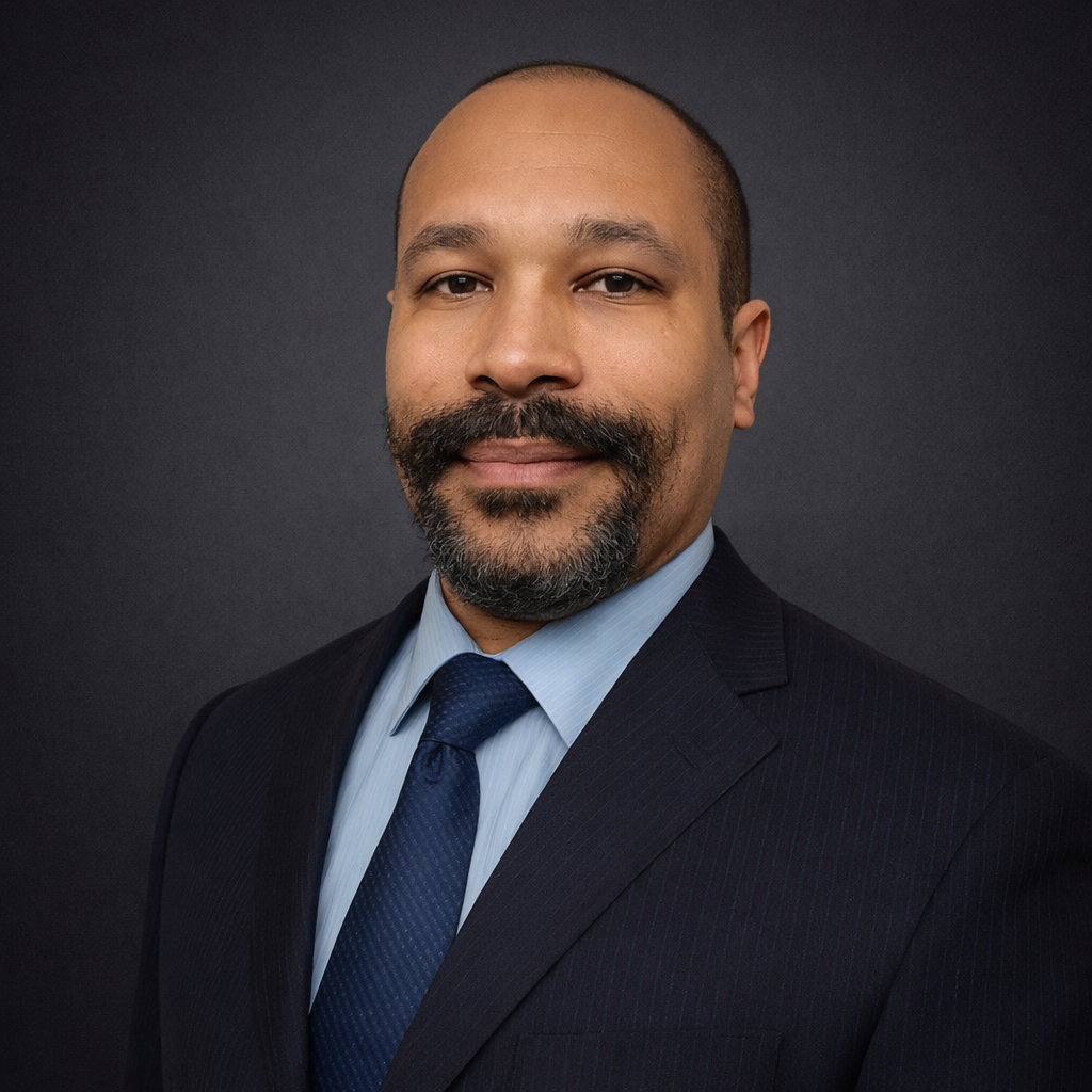 Headshot of a man in a dark suit, light blue shirt, and blue tie, against a dark background.