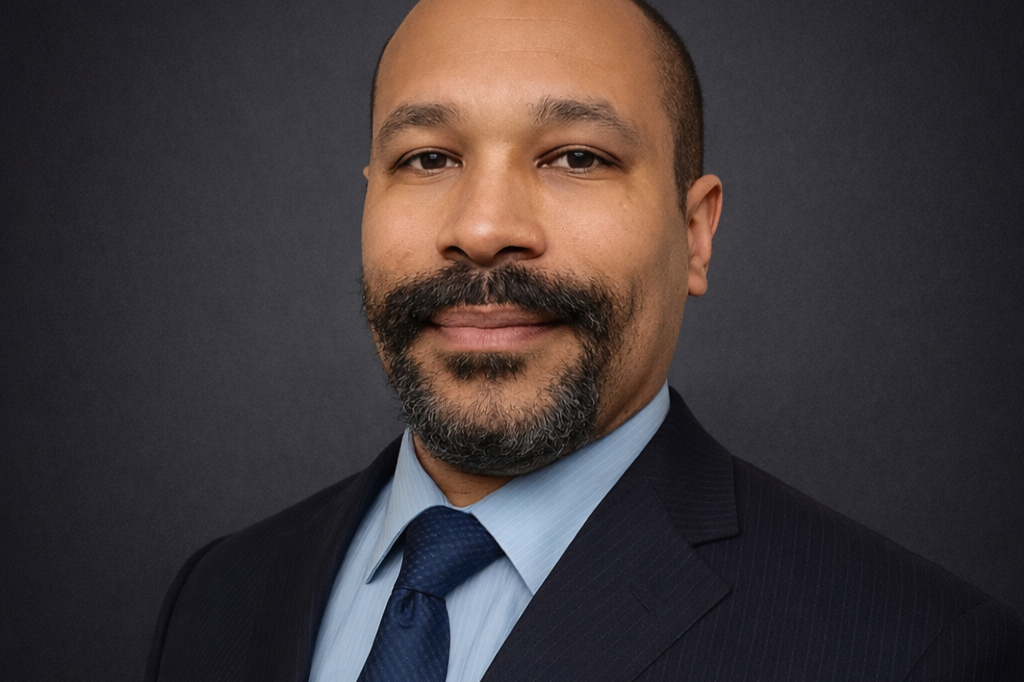 A professional man with a mustache and goatee, wearing a dark suit, light blue shirt, and navy tie, posing against a dark background.