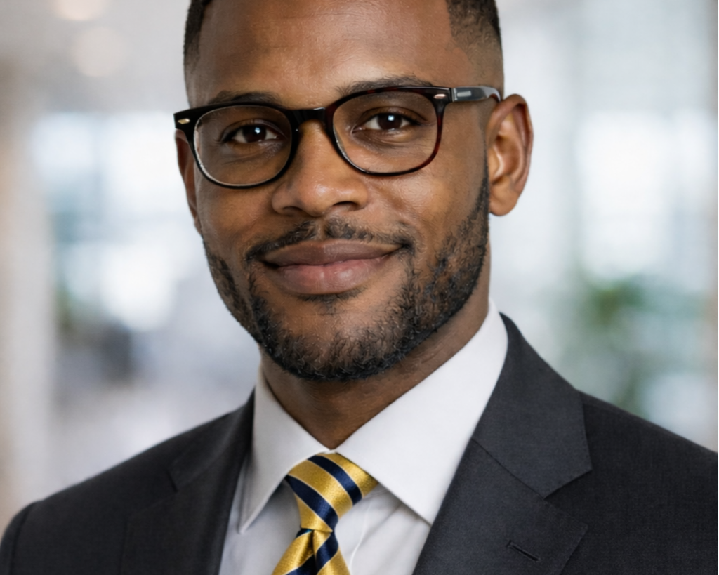 A man wearing glasses, a white dress shirt, a dark suit, and a yellow and blue striped tie, smiling indoors with a blurred background.