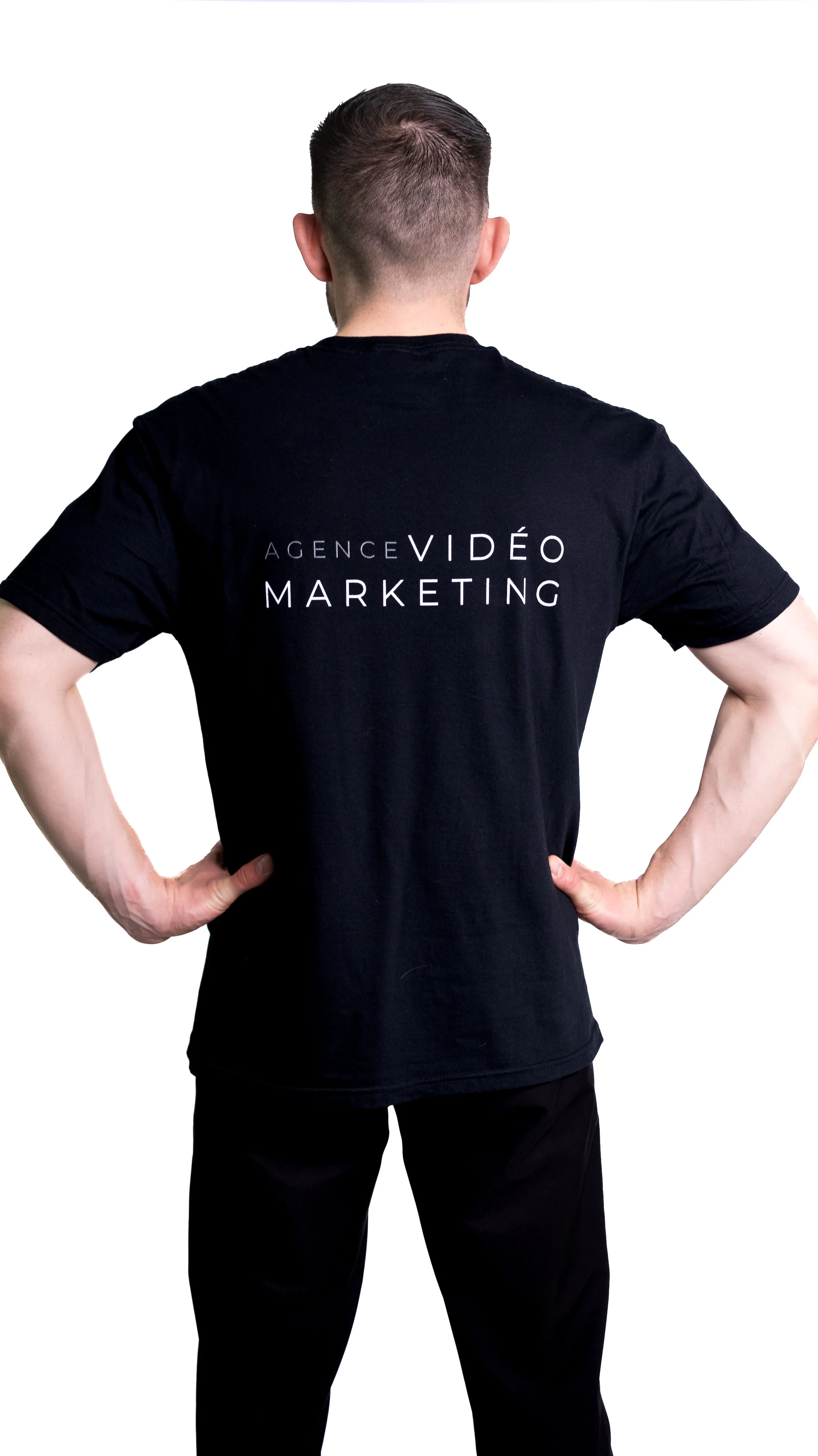 Homme vu de dos portant un t-shirt noir avec le texte 'Agence Vidéo Marketing' écrit dessus, debout avec les mains sur les hanches, contre un fond blanc.