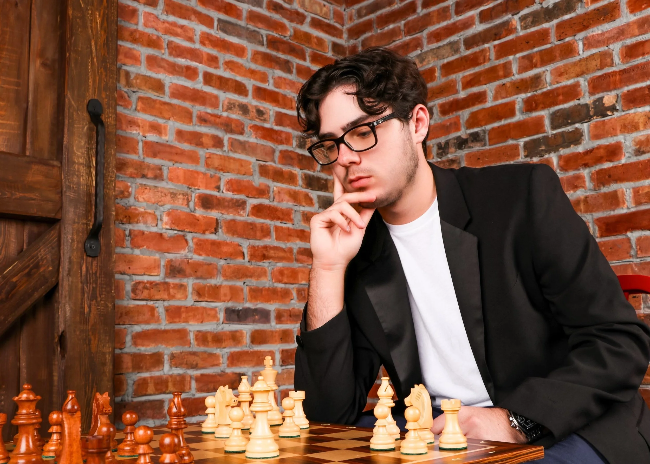 A young man with glasses, wearing a black blazer and white T-shirt, deeply focused on a chess game in a room with a brick wall background.