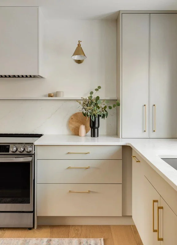 Modern kitchen with white cabinets, a white marble countertop, a black vase with green foliage, a wooden cutting board, and gold handles.