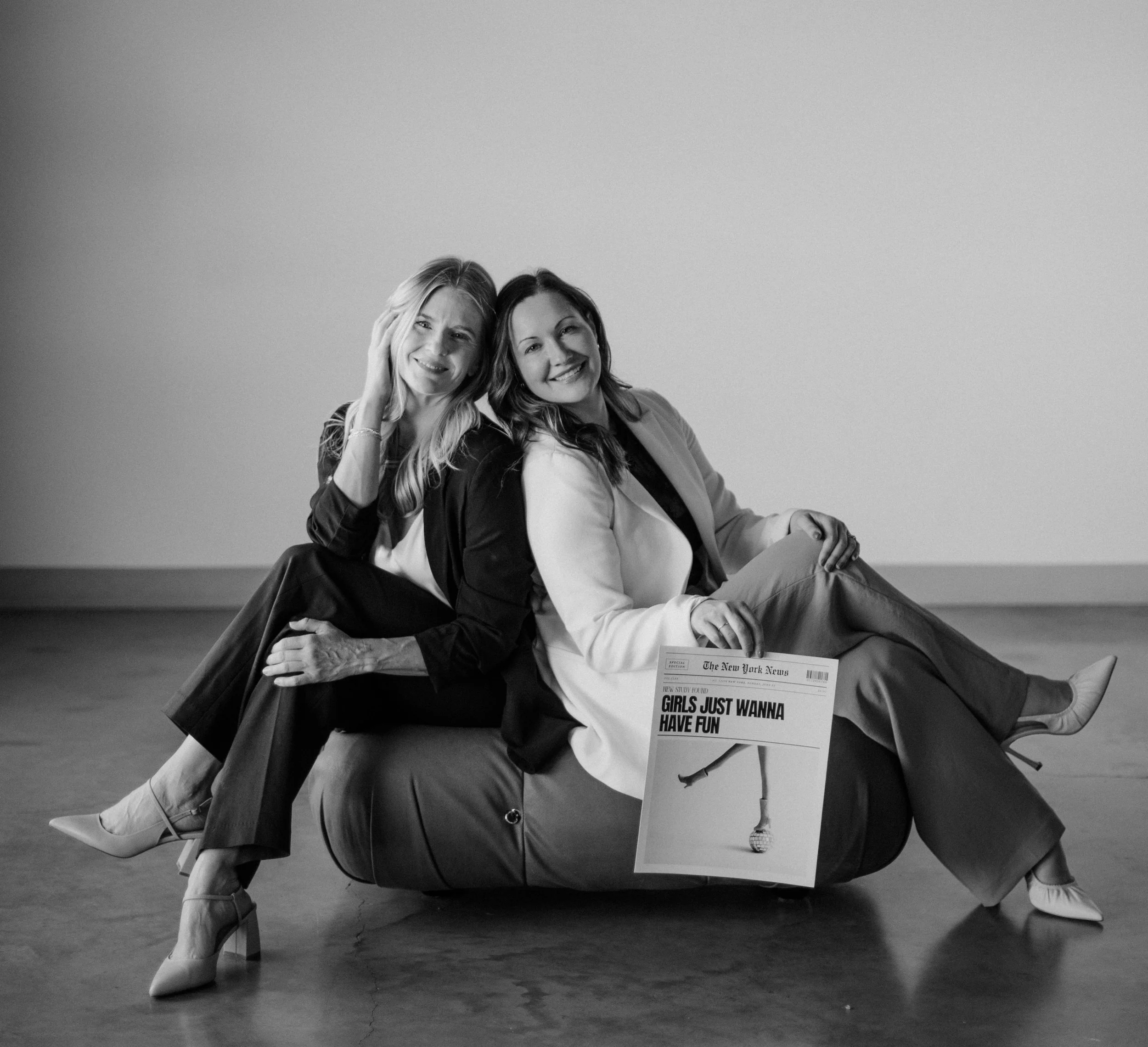 Two women sitting on a cushioned surface, smiling at the camera in a black-and-white photo. One woman is supporting the other, who is sitting sideways with legs crossed. They are holding a newspaper titled 'GIRLS JUST WANNA HAVE FUN' with a picture of a microphone on the cover.