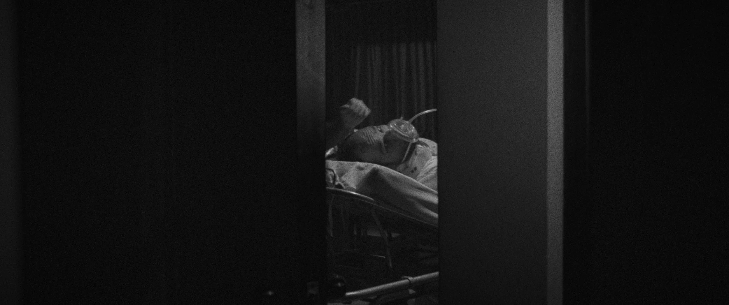 The Nation - Paciente en cama con oxígeno en una habitación oscura, visible a través de una rendija o puerta entreabierta