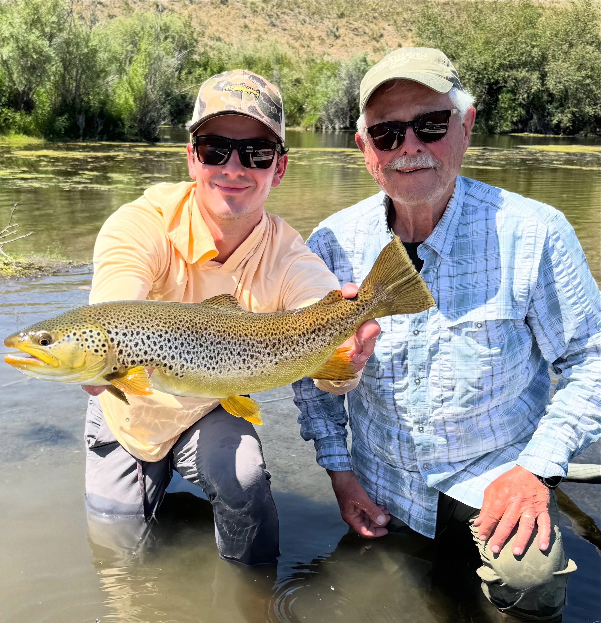 Two men standing in a shallow river holding a large rainbow trout fish, with a background of trees and a hilly landscape, both wearing sunglasses and hats.