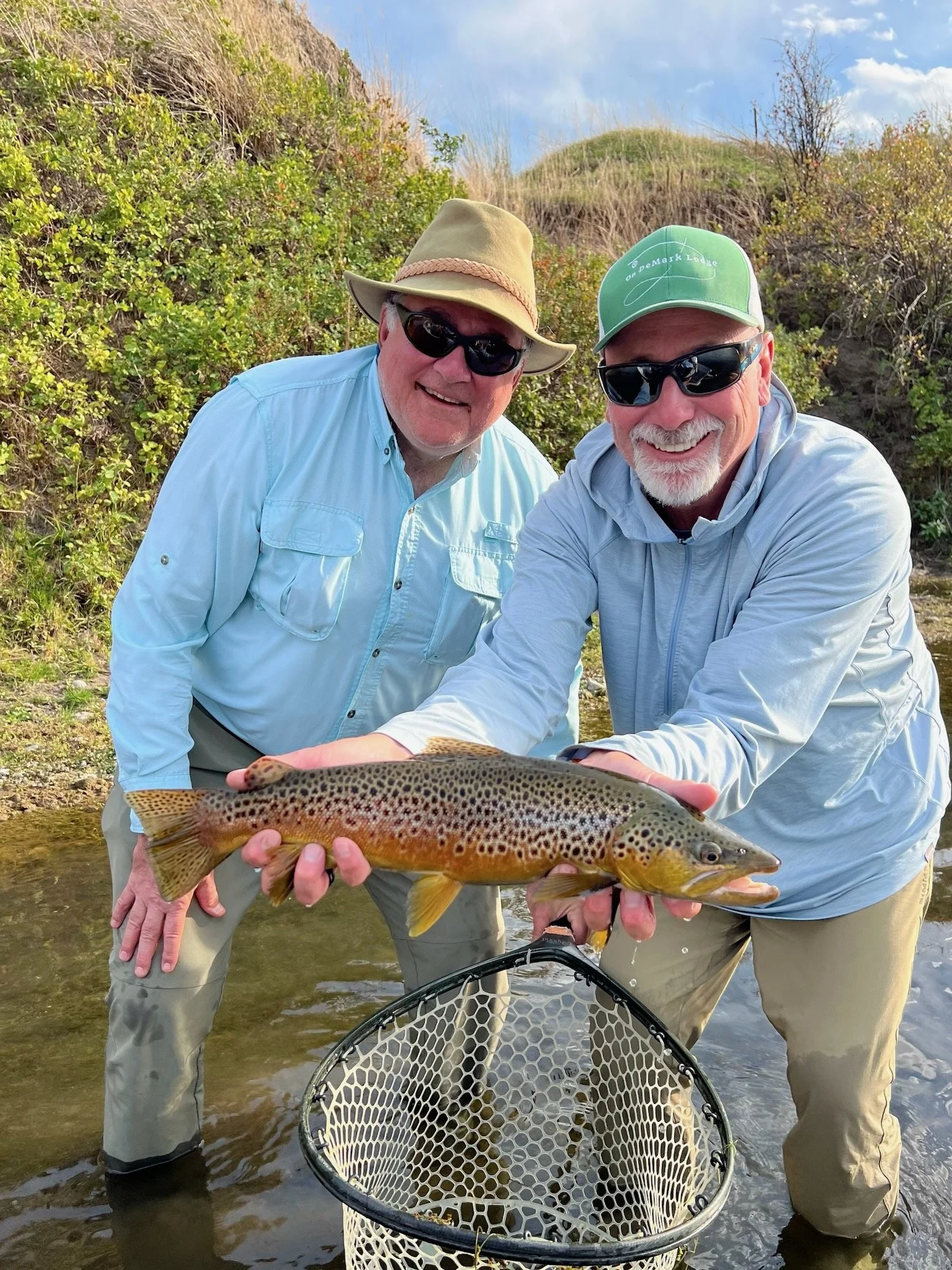 Two men standing in shallow water holding a large brown trout above a fishing net.