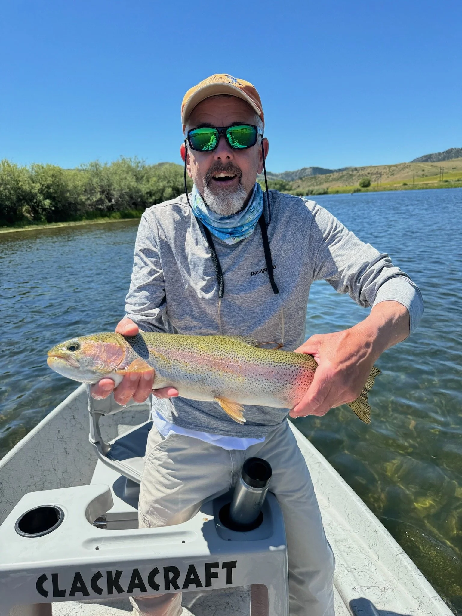 Man holding a rainbow trout while sitting in a boat on a river, wearing sunglasses and a beige cap, with a scenic landscape in the background.