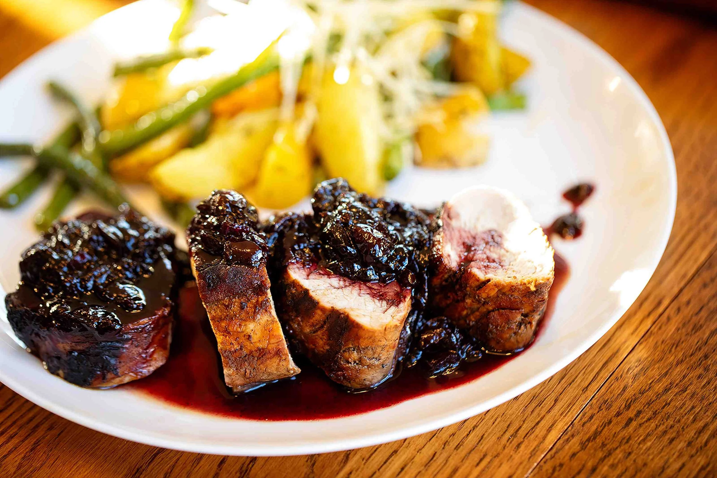 Sliced meat with dark berry sauce on a white plate, accompanied by potatoes and green beans.