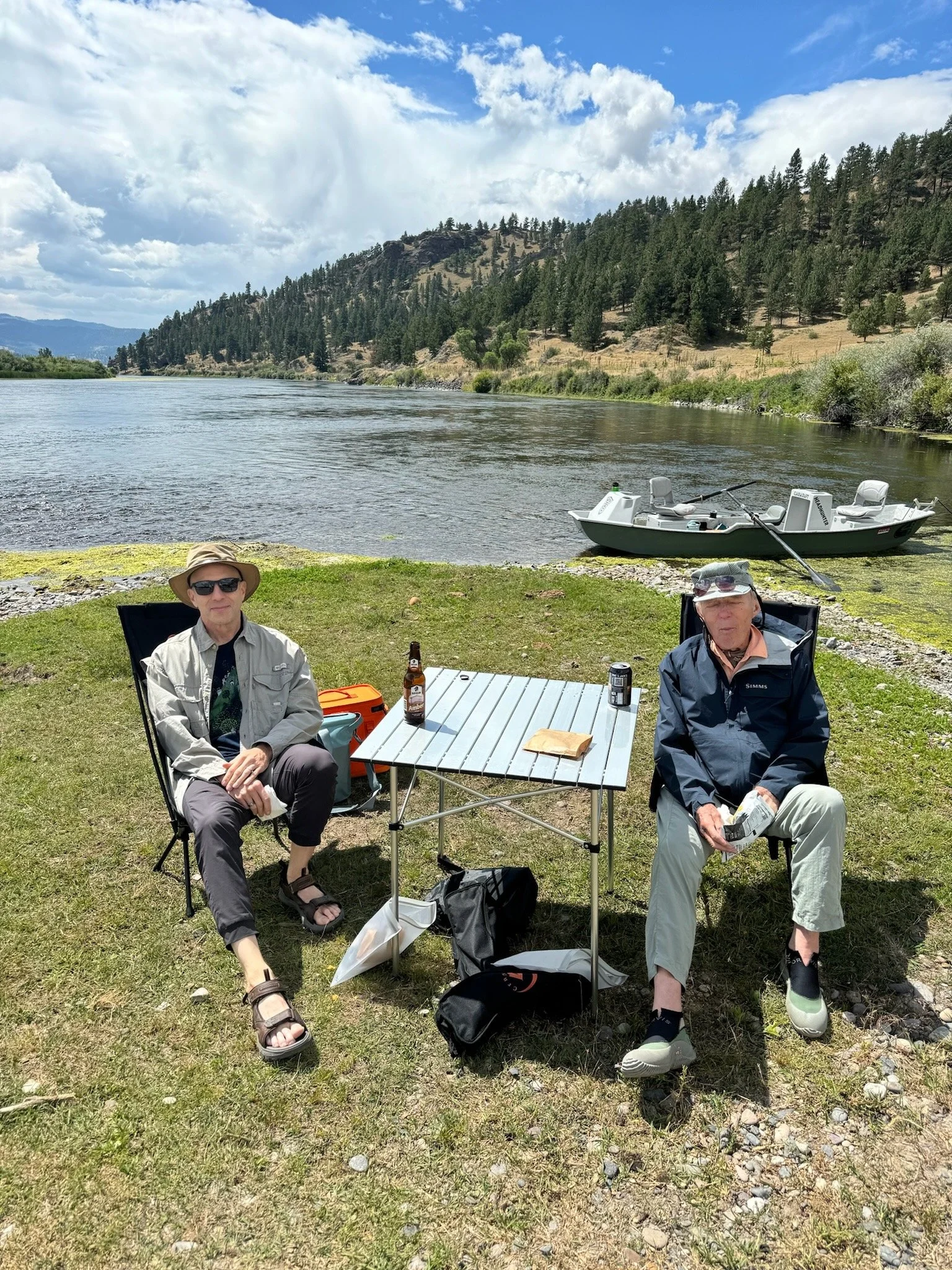 Two men sitting by a lakeside with a table, drinks, and a boat in the background, surrounded by mountainous landscape and forest.