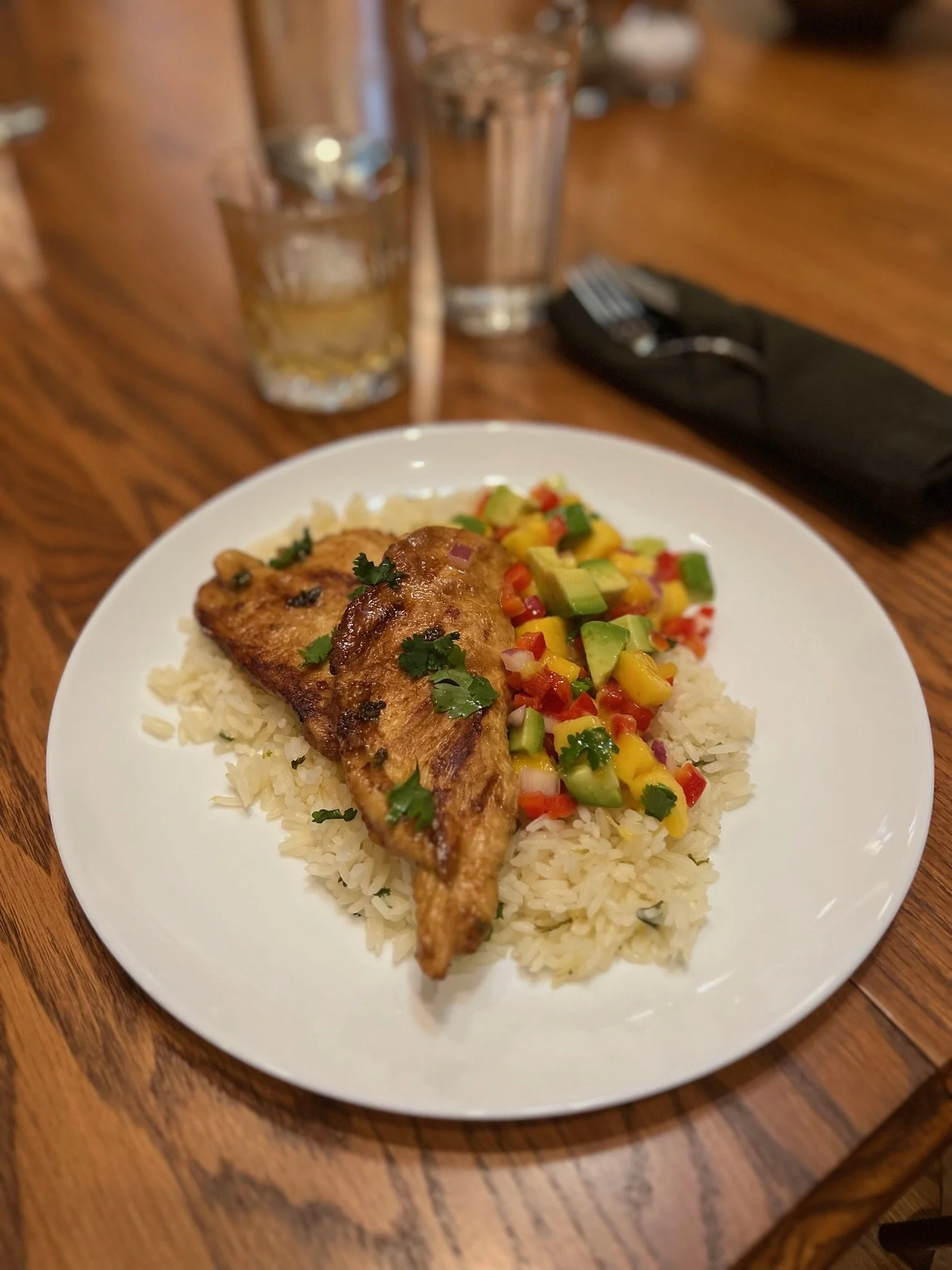 Grilled chicken on rice with mango, avocado, red bell pepper, and cilantro salsa on a white plate.