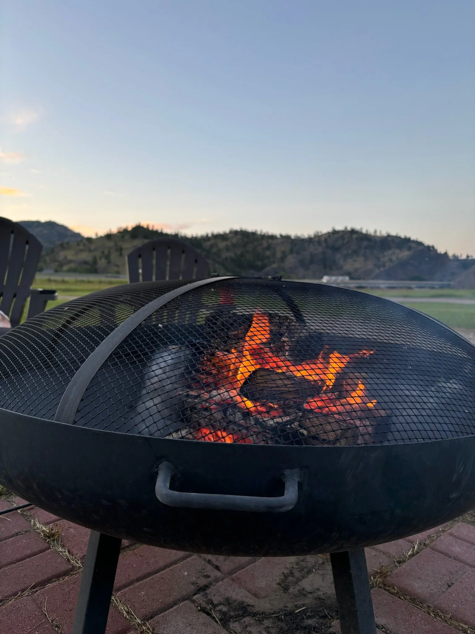 Outdoor fire pit with burning logs, surrounded by Adirondack chairs, against a backdrop of mountains and a clear evening sky.