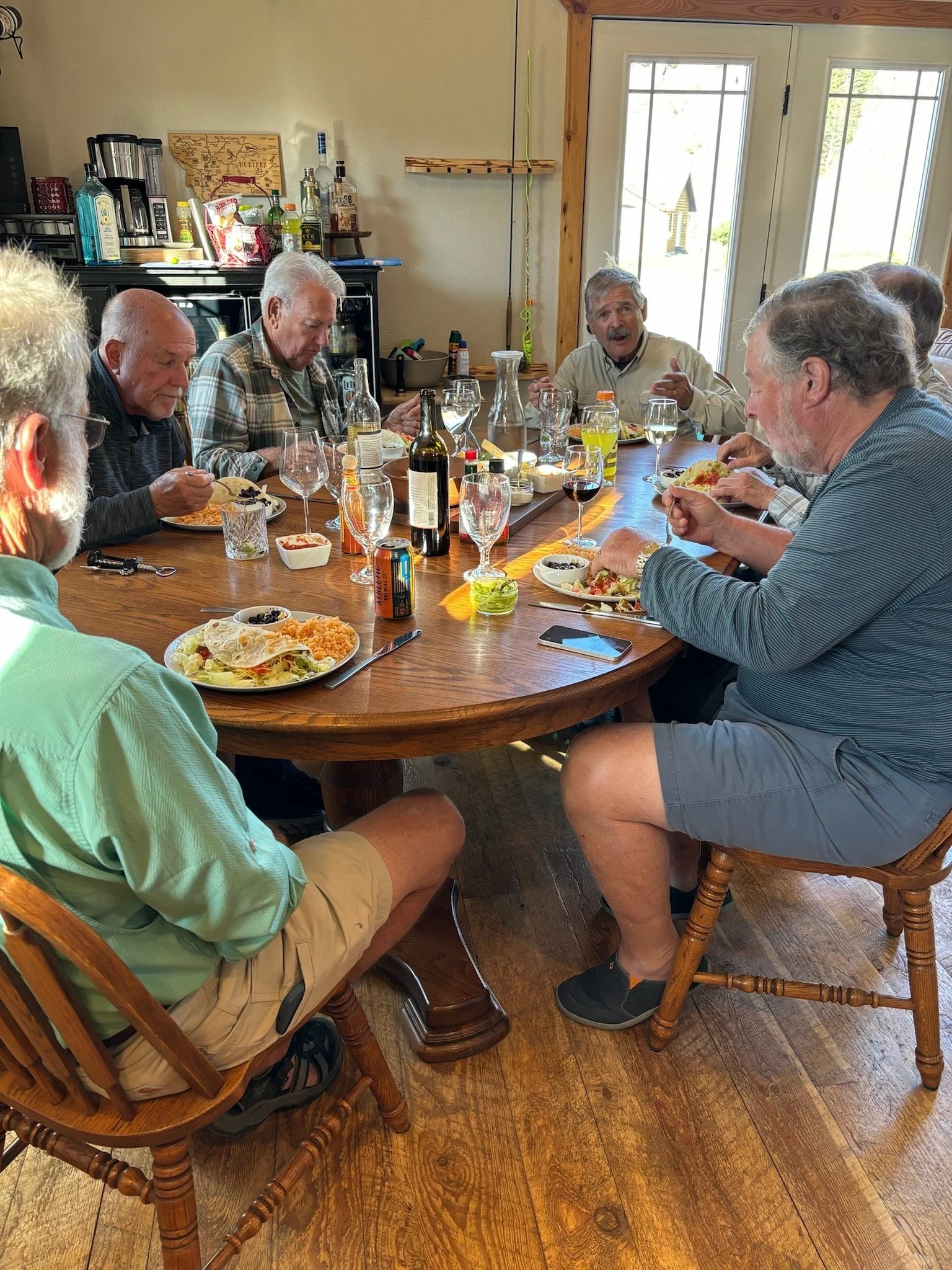 A group of older men sitting around a wooden dining table, sharing a meal. There are plates of food, wine glasses, and various bottles on the table. The room has wooden floors and glass pane doors in the background.