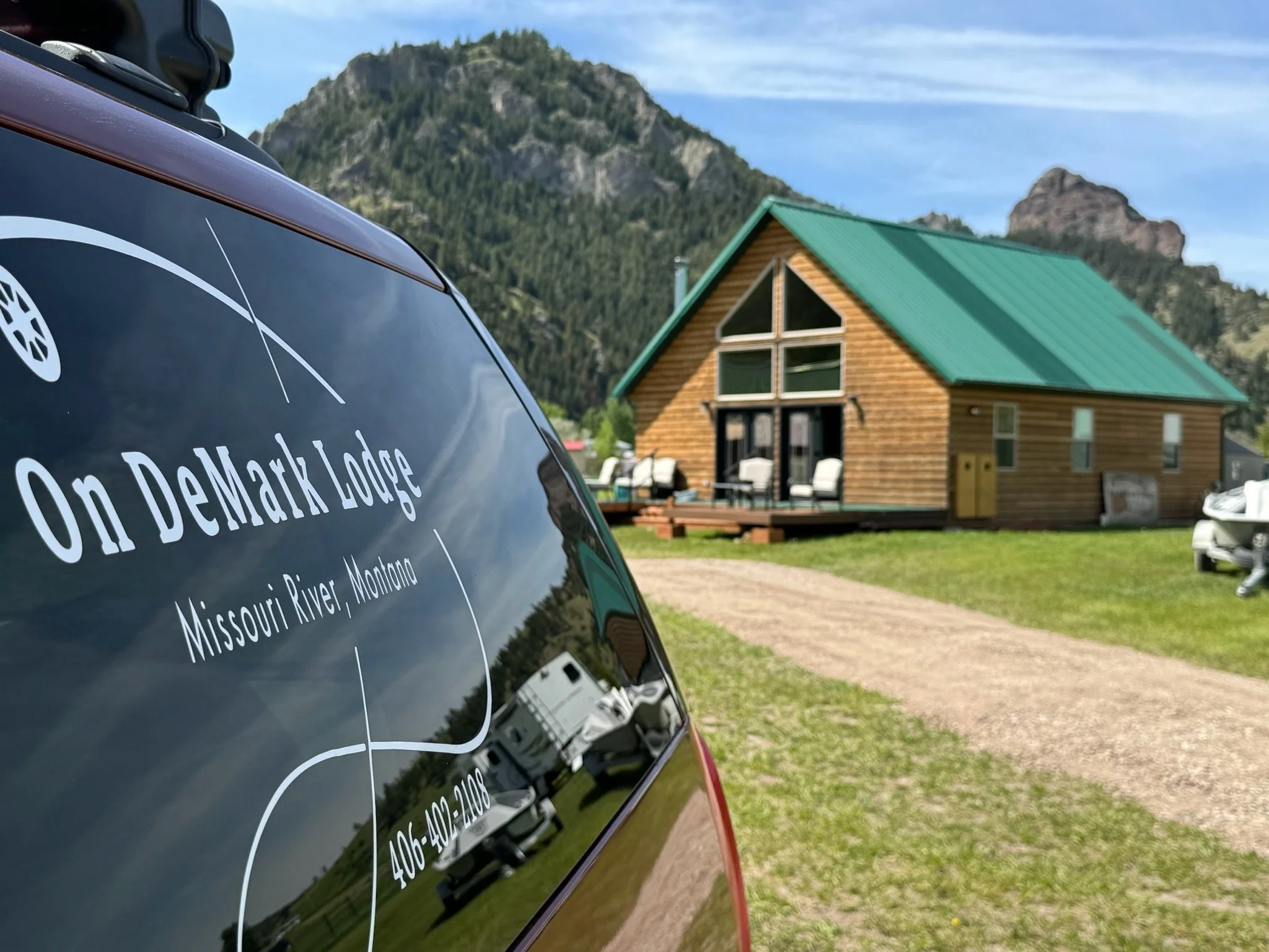 Car window with "On DeMark Lodge, Missouri River, Montana" decal, cabin with green roof and mountains in the background.