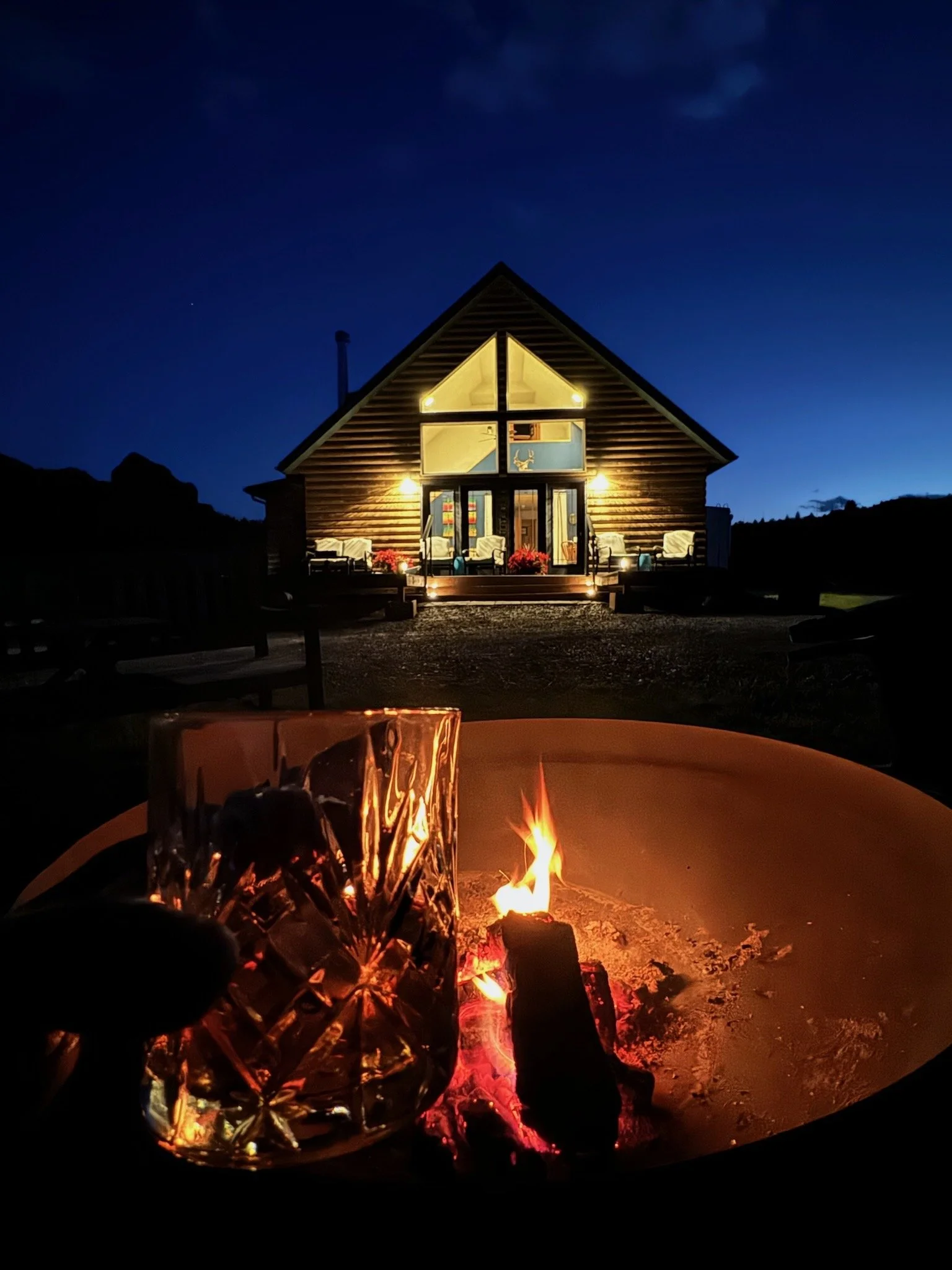 A night scene featuring a cozy cabin with a steeply pitched roof, warmly lit from within. In the foreground, a fire pit is burning, and a hand holds a glass of whiskey in front of the fire.
