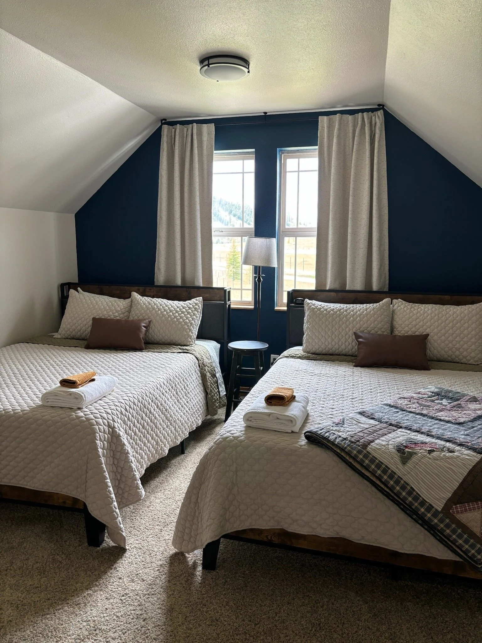 A bedroom with two neatly made beds, featuring light-colored quilts and stacked towels. A bedside lamp is positioned between the beds under two windows with curtains. The walls are partly painted blue, and the room has a sloped ceiling.