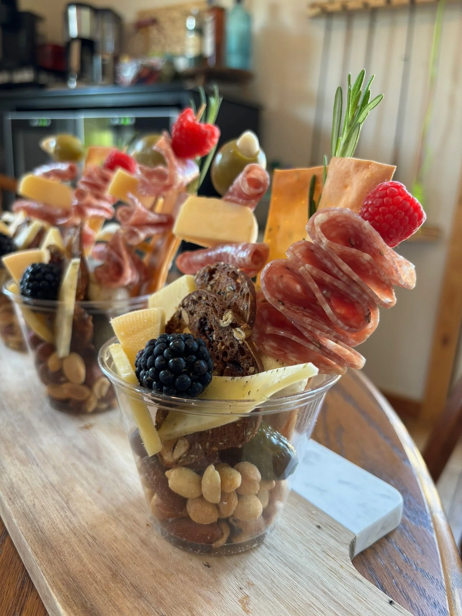 Charcuterie cups with meats, cheeses, nuts, blackberries, raspberries, and pickles on a wooden board.