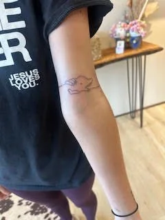 A person's arm with a small line drawing tattoo of a whale on the inner forearm, and a black hair tie on the wrist. The background shows a wooden table with a small bouquet of flowers and some decor.