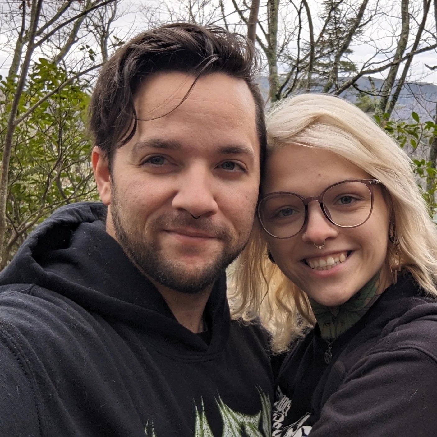 A young man and woman taking a selfie outdoors with trees and a cloudy sky in the background.