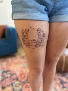 Tattoo of a horseshoe with flowers and ribbons on a person's thigh, wearing denim shorts.