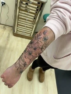 A person's arm with various colorful tattoos, including flowers and geometric designs, wearing a light pink sweatshirt.