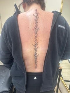Back tattoo of a vertical floral vine design running down the spine, partly covered by a black hoodie.