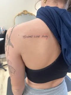Tattoo reading 'Strength Love Andy' on a person's shoulder. The person is wearing a black top and a blue hoodie with a tattoo on their arm.