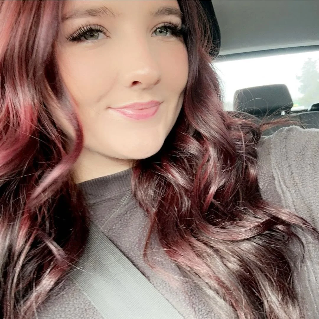 Close-up of a smiling woman with long, wavy, reddish-brown hair sitting in a car.