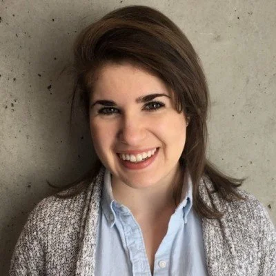 Smiling young woman with brown hair wearing a light blue button-down shirt and gray knit cardigan, posed against a concrete wall.