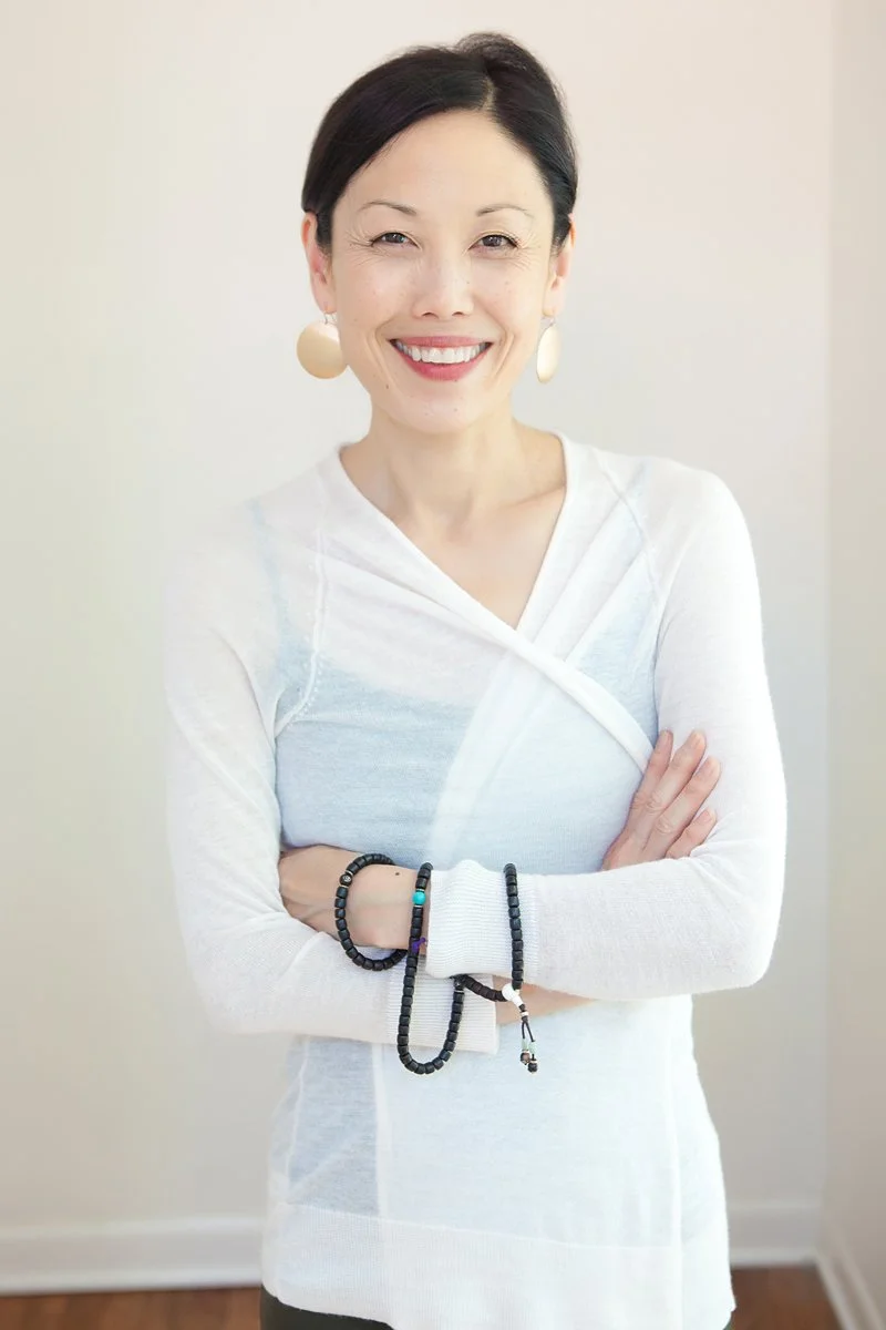 Smiling woman standing with arms crossed, wearing a white wrap top, large round earrings and beaded bracelets, against a pale wall.