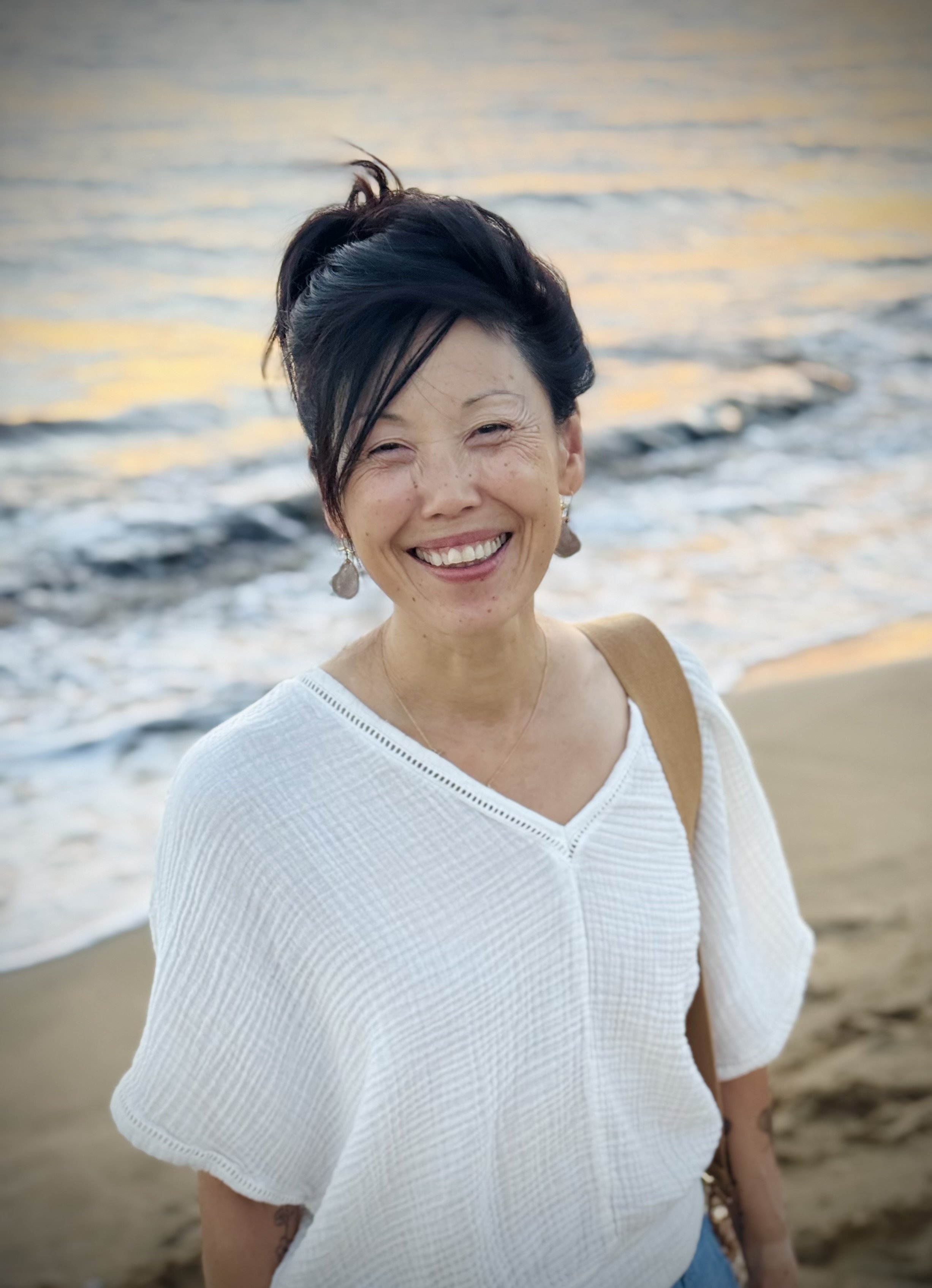 Smiling woman standing on a beach at sunset, wearing a white textured top and dangling earrings, with waves softly blurred behind her.