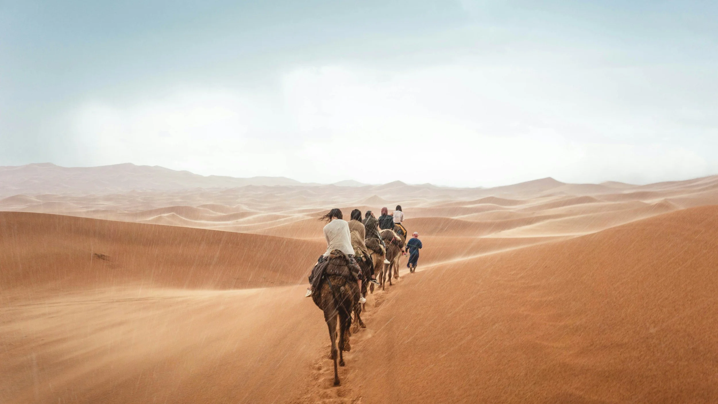 A group of people riding camels in a desert landscape with rolling sand dunes.