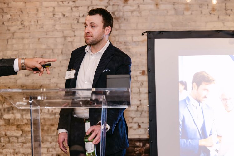 Speaker at a clear lectern beside a presentation screen in a brick-walled venue