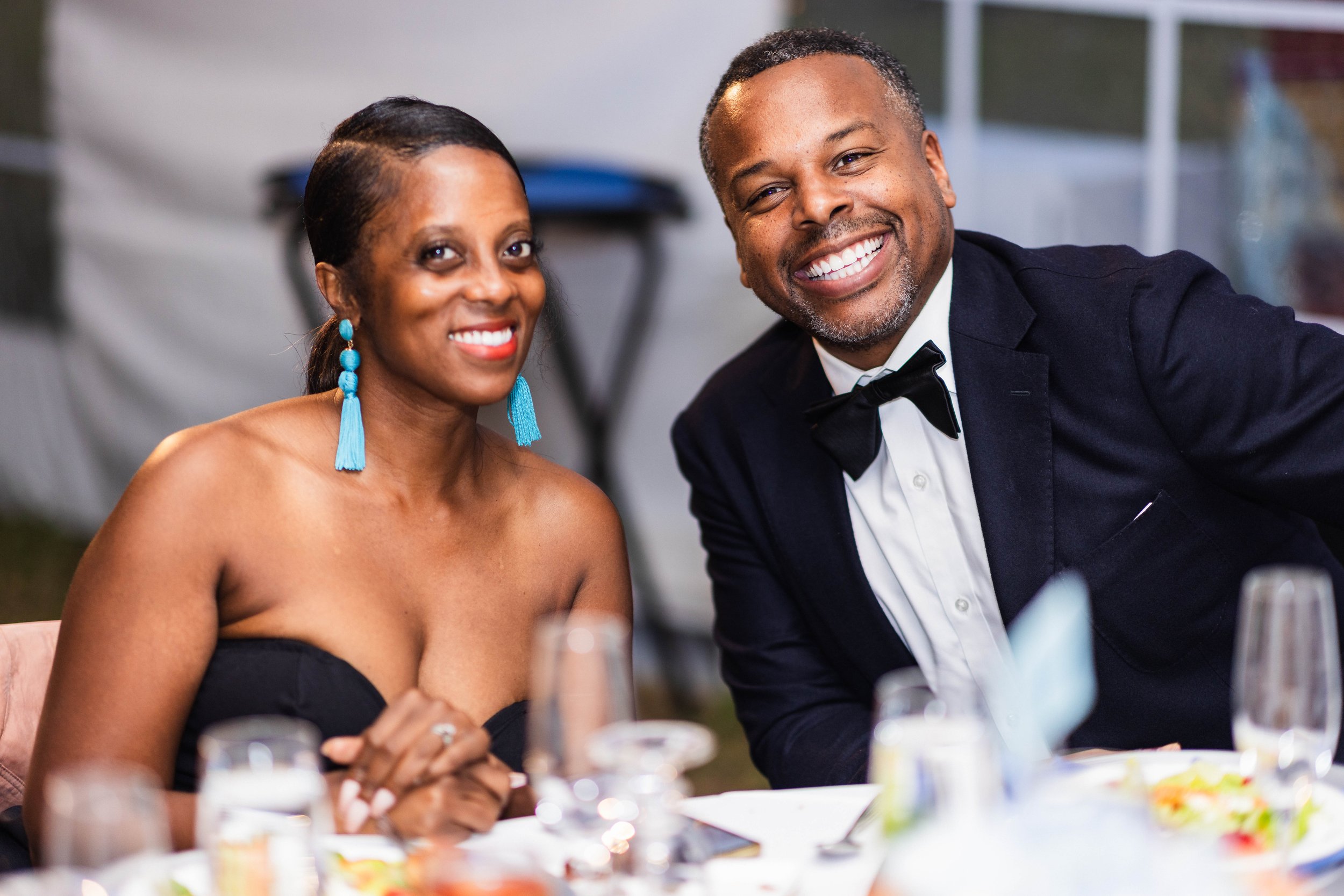 A smiling woman in a black strapless dress with blue earrings and a smiling man in a tuxedo at a formal gathering.