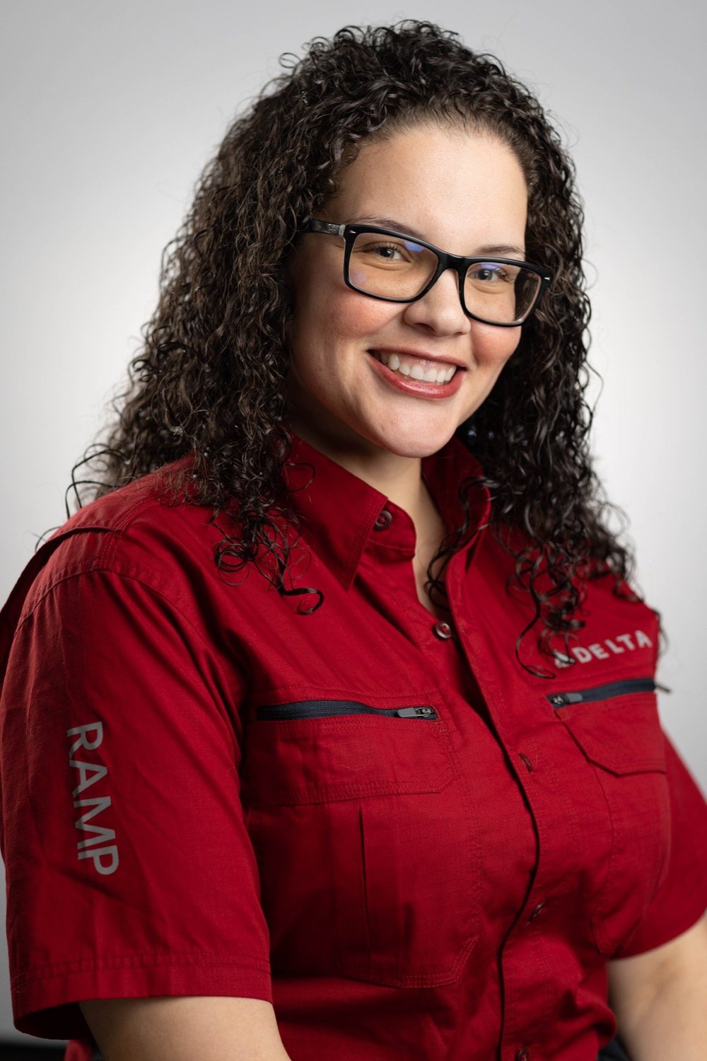 Professional portrait of a smiling woman with glasses in a red work shirt against a light gray backdrop