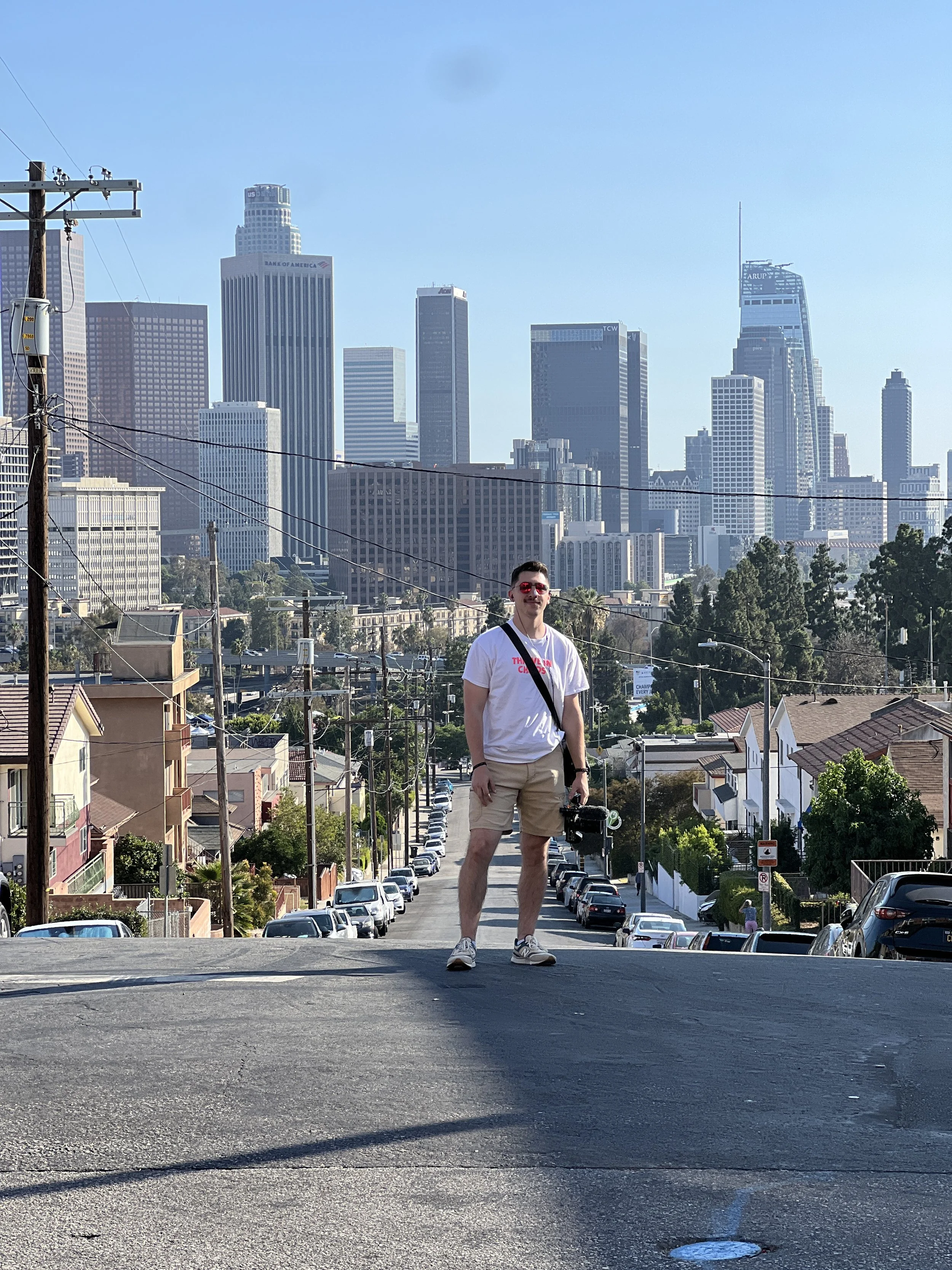 A zoomed landscape photo of Nico with his camera being the main subject and the city-scape of Los Angeles behind him. 