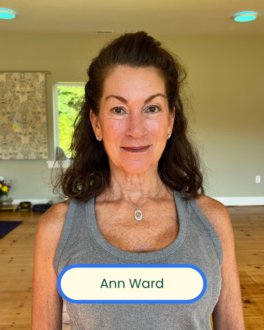 A woman with dark brown hair wearing a gray tank top and jewelry, standing indoors with a background of a green outdoor view, wooden floor, and minimal decor.