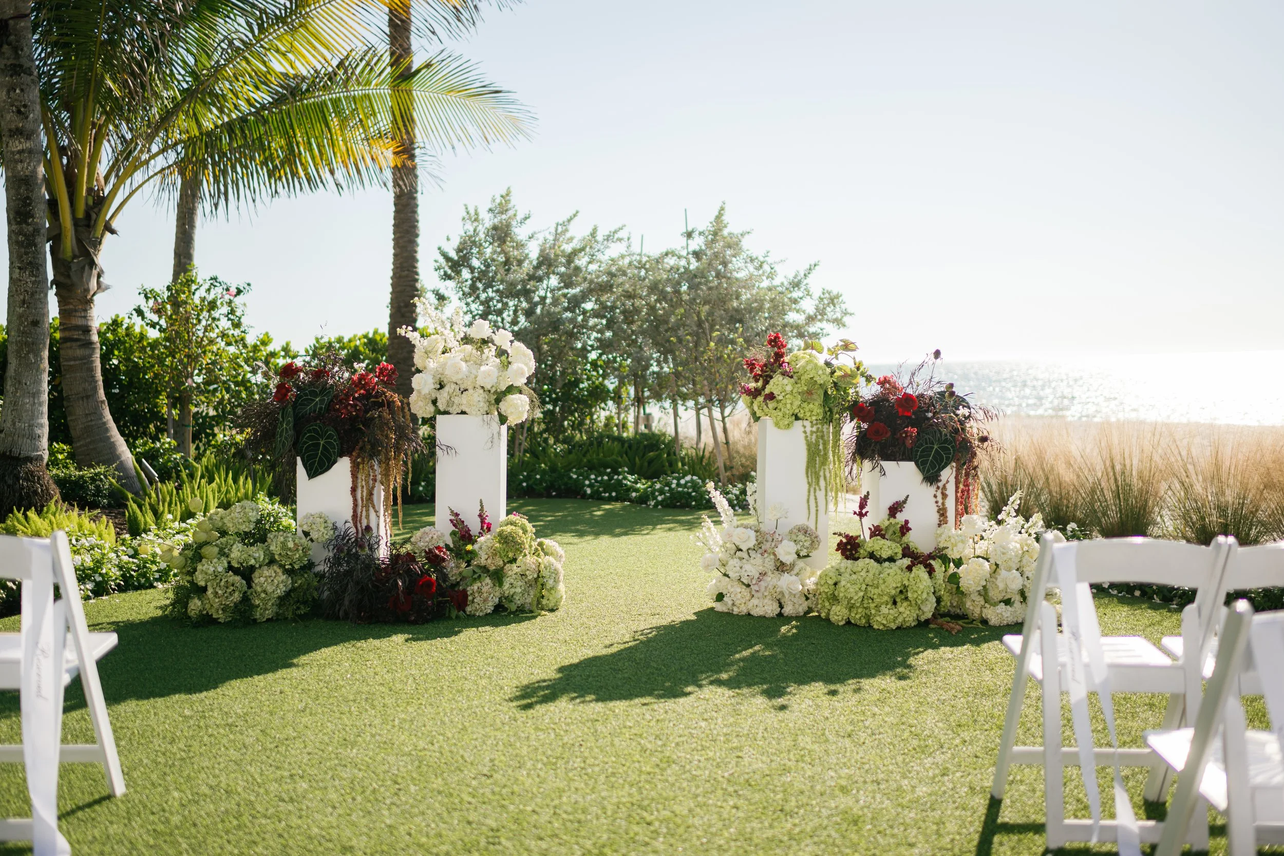 Beachside outdoor wedding setup with white chairs, floral arrangements with white and dark red flowers, and lush greenery near the ocean with palm trees and tall grasses in the background.