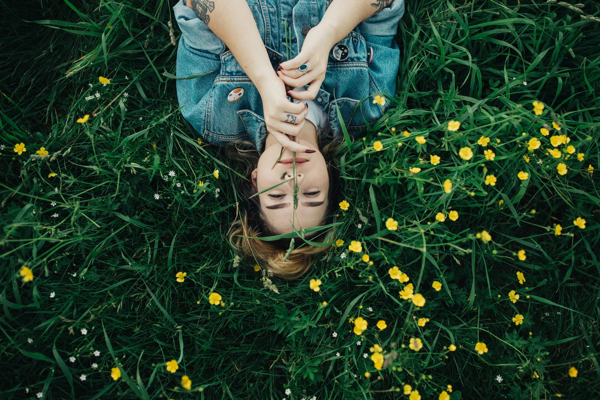 A woman lying in a grassy field surrounded by yellow and white wildflowers, with her eyes closed and fingers near her lips, wearing a denim jacket and rings.