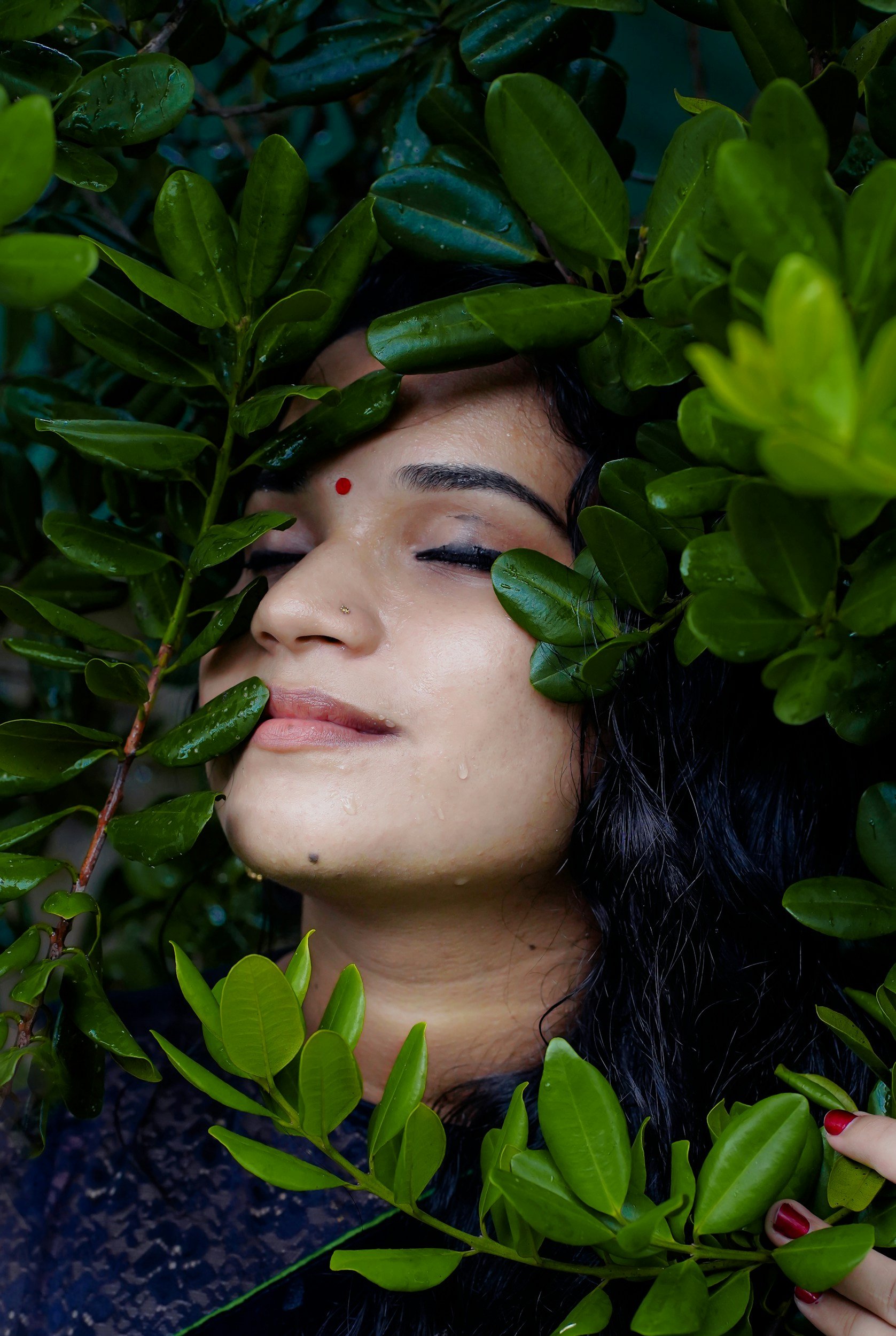 A woman with black hair and light brown skin is surrounded by lush green leaves, with her eyes closed and a peaceful expression.