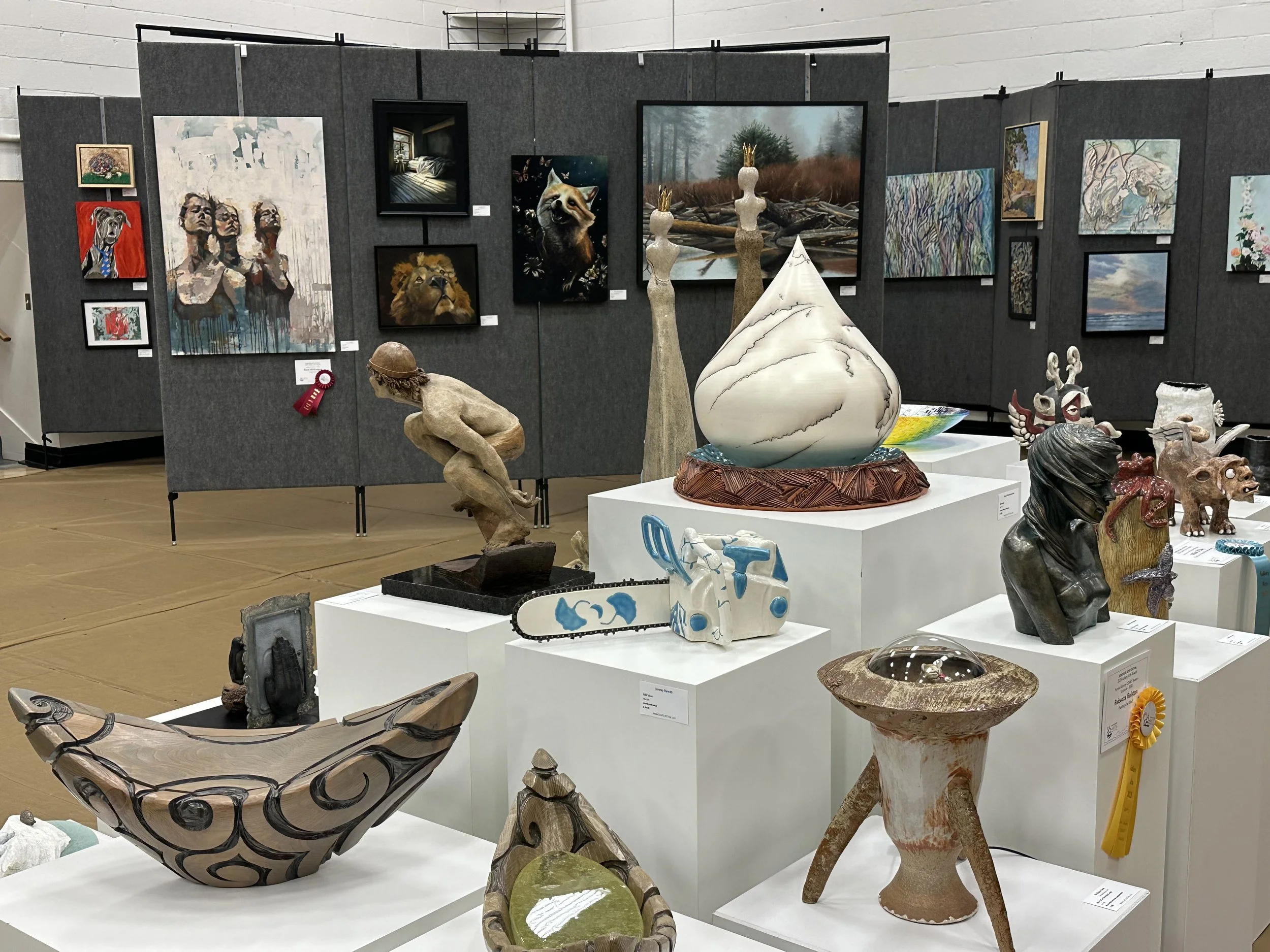 Art gallery with various paintings, sculptures, and crafts displayed on white pedestals and gray walls.