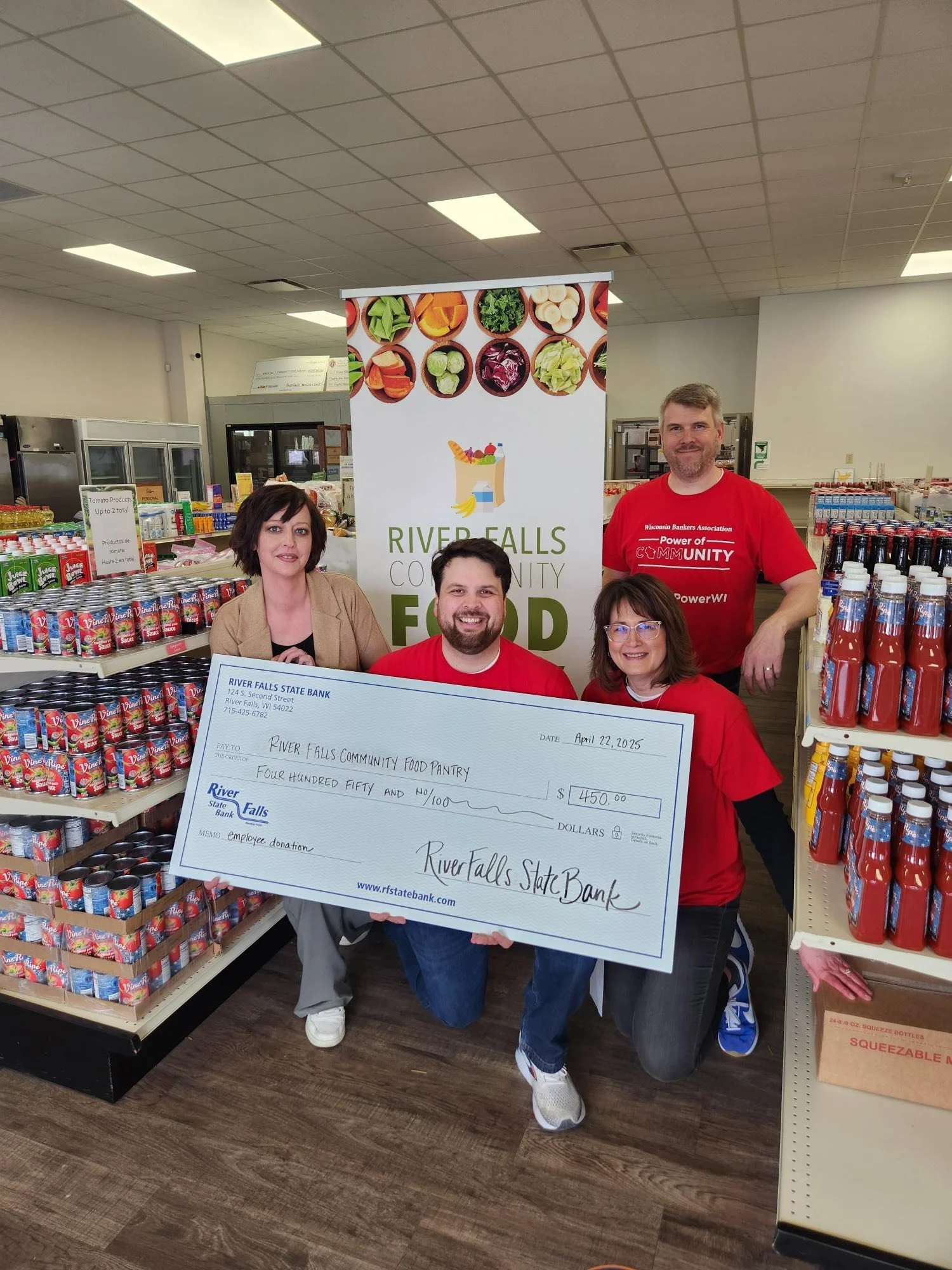 Four people, two sitting and two standing, hold a large donation check inside a food pantry. The check is for $450 from River Falls State Bank to River Falls Community Food Pantry. Shelves of canned goods and a banner with the pantry name are visible in the background, indicating a charitable event.