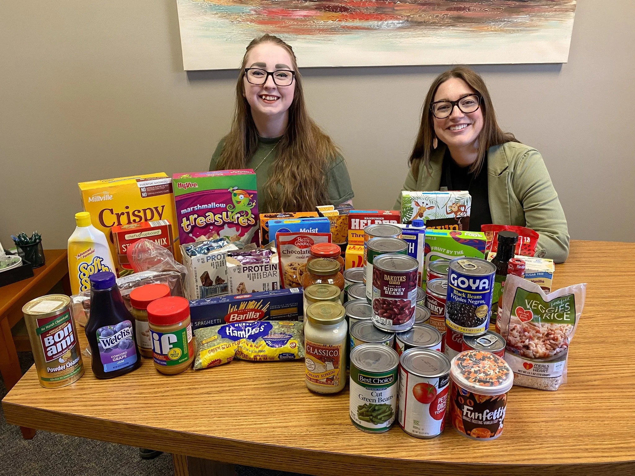 Two people sitting behind a table filled with various food products including cereal, canned goods, pasta, and snacks.